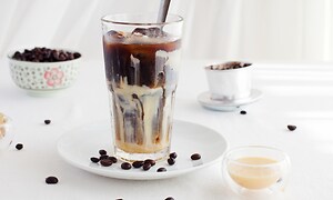 SDA - OBH Nordica - Et højt glas fyldt med iskaffe på et hvidt bord med kaffebønner spredt rundt omkring